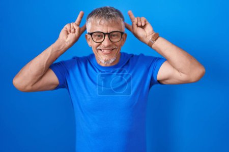 Téléchargez les photos : Homme hispanique avec les cheveux gris debout sur fond bleu posant drôle et fou avec les doigts sur la tête comme des oreilles de lapin, souriant gai - en image libre de droit