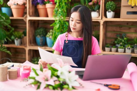 Foto de Young chinese woman florist using laptop reading document at flower shop - Imagen libre de derechos