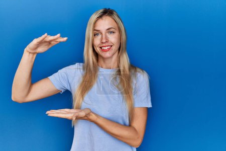 Photo for Beautiful blonde woman wearing casual t shirt over blue background gesturing with hands showing big and large size sign, measure symbol. smiling looking at the camera. measuring concept. - Royalty Free Image