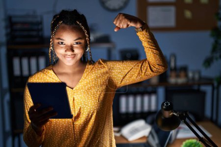 Photo for African american woman with braids working at the office at night with tablet strong person showing arm muscle, confident and proud of power - Royalty Free Image