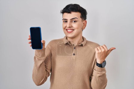 Photo for Non binary person holding smartphone showing blank screen pointing thumb up to the side smiling happy with open mouth - Royalty Free Image