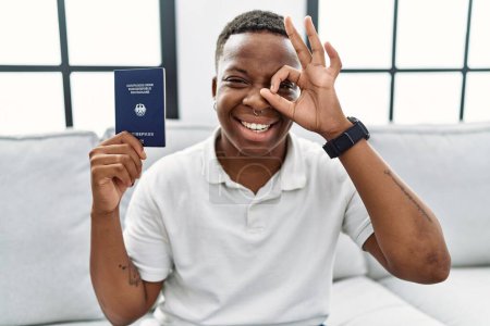 Foto de Joven africano portador de pasaporte deutschland sonriendo feliz haciendo señal bien con la mano en el ojo mirando a través de los dedos - Imagen libre de derechos