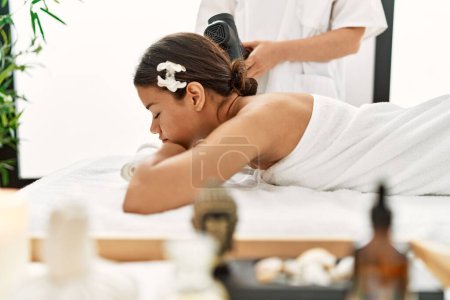 Photo for Young latin woman relaxed having back massage with percussion pistol at beauty center - Royalty Free Image