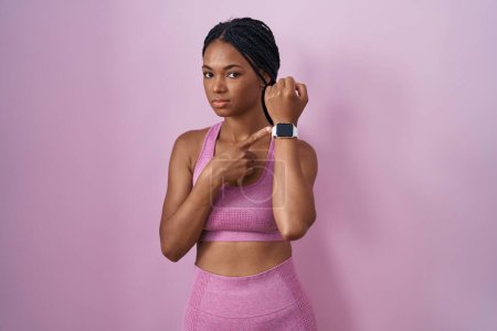 Téléchargez les photos : African american woman with braids wearing sportswear over pink background in hurry pointing to watch time, impatience, looking at the camera with relaxed expression - en image libre de droit