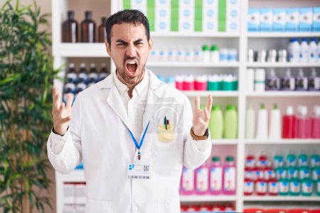 Foto de Handsome hispanic man working at pharmacy drugstore crazy and mad shouting and yelling with aggressive expression and arms raised. frustration concept. - Imagen libre de derechos