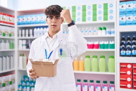 Foto de Adolescente hispano trabajando en farmacia en una caja de espera con pastillas molesto y frustrado gritando de rabia, gritando loco de rabia y levantando la mano - Imagen libre de derechos