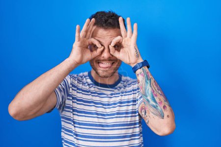 Foto de Joven hombre hispano de pie sobre fondo azul haciendo un buen gesto como binoculares sacando la lengua, ojos mirando a través de los dedos. expresión loca. - Imagen libre de derechos