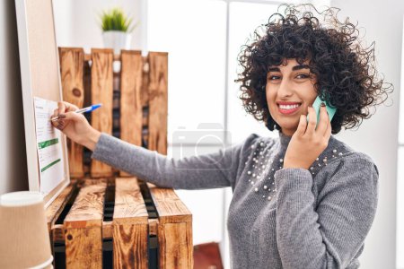 Foto de Young middle east woman business worker talking on smartphone writing on cork board at office - Imagen libre de derechos