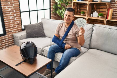 Foto de Joven hispano con el brazo en la honda sosteniendo billetes de dólares de EE.UU. guiñando el ojo mirando a la cámara con expresión sexy, cara alegre y feliz. - Imagen libre de derechos