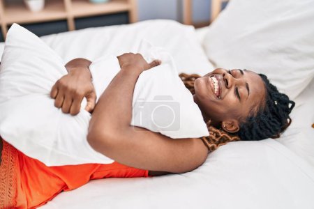 Téléchargez les photos : Femme afro-américaine étreignant oreiller couché sur le lit dans la chambre - en image libre de droit