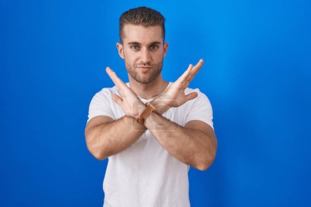 Photo for Young caucasian man standing over blue background rejection expression crossing arms doing negative sign, angry face - Royalty Free Image