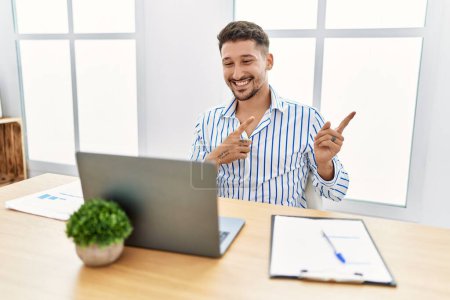 Foto de Joven hombre guapo con barba trabajando en la oficina usando computadora portátil sonriendo y mirando a la cámara apuntando con dos manos y dedos hacia un lado. - Imagen libre de derechos