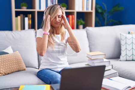 Foto de Mujer rubia joven estudiando el uso de ordenador portátil en casa con la mano en la cabeza, dolor de cabeza porque el estrés. padecer migraña. - Imagen libre de derechos