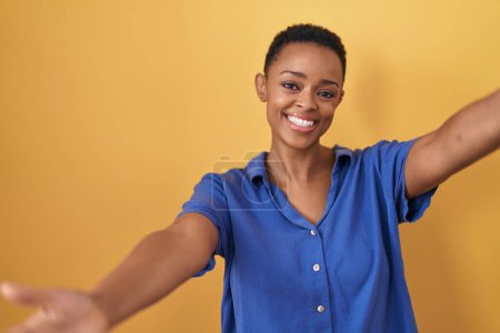 Téléchargez les photos : Femme afro-américaine debout sur fond jaune regardant la caméra souriant à bras ouverts pour un câlin. expression joyeuse embrassant le bonheur. - en image libre de droit