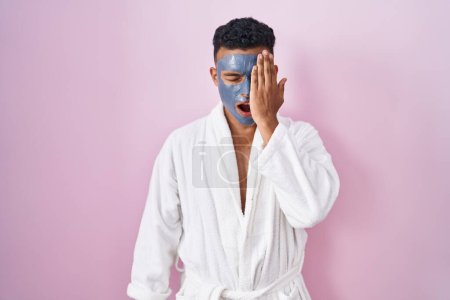 Photo for Young hispanic man wearing beauty face mask and bath robe yawning tired covering half face, eye and mouth with hand. face hurts in pain. - Royalty Free Image