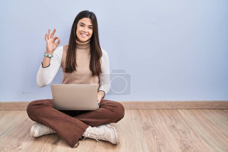 Foto de Mujer morena joven trabajando con computadora portátil sentado en el suelo sonriendo positiva haciendo signo de ok con la mano y los dedos. expresión exitosa. - Imagen libre de derechos
