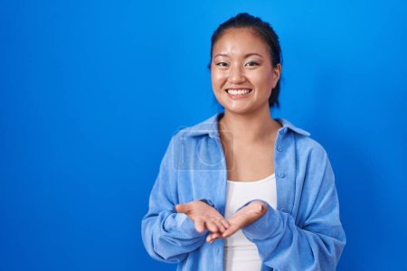 Téléchargez les photos : Asiatique jeune femme debout sur fond bleu souriant avec les mains paumes ensemble recevoir ou donner geste. maintien et protection - en image libre de droit