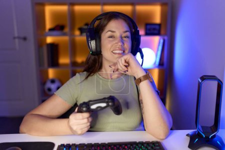 Photo for Beautiful brunette woman playing video games wearing headphones with hand on chin thinking about question, pensive expression. smiling and thoughtful face. doubt concept. - Royalty Free Image