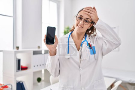Photo for Young hispanic doctor woman holding smartphone showing screen stressed and frustrated with hand on head, surprised and angry face - Royalty Free Image