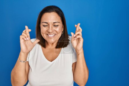 Foto de Mujer madura hispana de pie sobre fondo azul gesto dedo cruzado sonriendo con esperanza y los ojos cerrados. suerte y concepto supersticioso. - Imagen libre de derechos