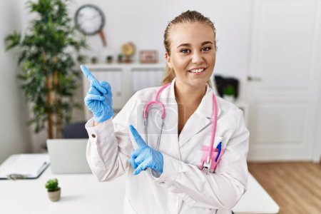 Photo for Young caucasian woman wearing doctor uniform and stethoscope at the clinic smiling and looking at the camera pointing with two hands and fingers to the side. - Royalty Free Image
