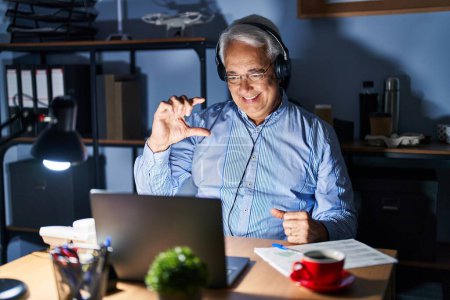 Photo for Hispanic senior man wearing call center agent headset at night smiling and confident gesturing with hand doing small size sign with fingers looking and the camera. measure concept. - Royalty Free Image
