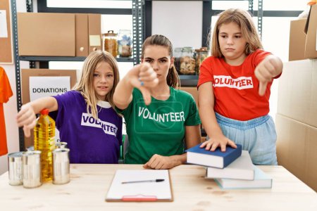 Foto de Young woman with two daughters wearing volunteer t shirt at donations stand with angry face, negative sign showing dislike with thumbs down, rejection concept - Imagen libre de derechos