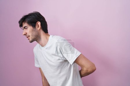 Téléchargez les photos : Jeune homme hispanique debout sur fond rose souffrant de maux de dos, touchant le dos avec la main, douleurs musculaires - en image libre de droit
