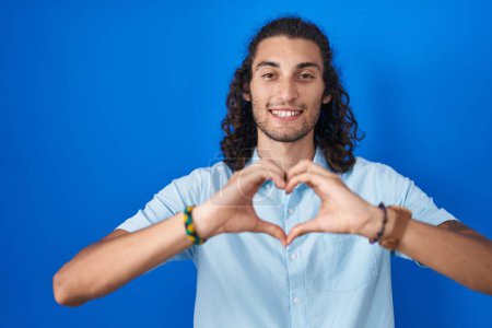 Photo for Young hispanic man standing over blue background smiling in love doing heart symbol shape with hands. romantic concept. - Royalty Free Image