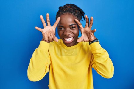 Photo for Beautiful black woman standing over blue background smiling funny doing claw gesture as cat, aggressive and sexy expression - Royalty Free Image