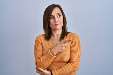 Photo for Middle age brunette woman standing wearing orange sweater pointing with hand finger to the side showing advertisement, serious and calm face - Royalty Free Image