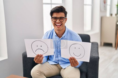Foto de Hombre hispano trabajando en la depresión sosteniendo triste a la emoción feliz papel sonriendo y riendo en voz alta porque broma loca divertida. - Imagen libre de derechos