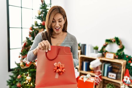 Photo for Young latin woman holding gift bag standing by christmas tree at home - Royalty Free Image