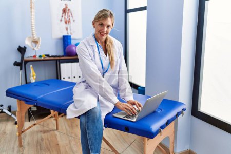 Téléchargez les photos : Young blonde woman wearing physiotherpist uniform using laptop at physiotherapy clinic - en image libre de droit
