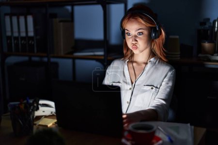 Téléchargez les photos : Jeune femme caucasienne travaillant au bureau la nuit gonflant les joues avec un visage drôle. bouche gonflée d'air, expression folle. - en image libre de droit