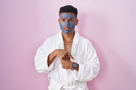Téléchargez les photos : Jeune homme hispanique portant un masque de beauté et peignoir de bain à la hâte pointant vers regarder le temps, l'impatience, bouleversé et en colère pour retard de délai - en image libre de droit