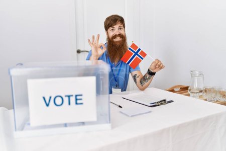 Foto de Hombre caucásico con barba larga en las elecciones de campaña política sosteniendo bandera noruega haciendo señal bien con los dedos, sonriendo gesto amistoso excelente símbolo - Imagen libre de derechos