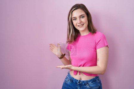 Téléchargez les photos : Blonde femme caucasienne debout sur fond rose invitant à entrer souriant naturel avec la main ouverte - en image libre de droit