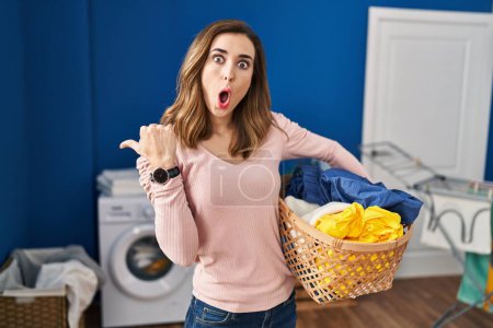 Photo for Young woman holding laundry basket surprised pointing with hand finger to the side, open mouth amazed expression. - Royalty Free Image