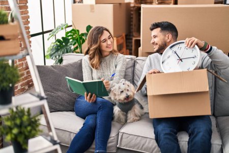 Photo for Man and woman unpacking box writing on notebook sitting on sofa with dog at new home - Royalty Free Image