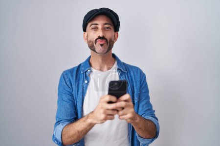 Téléchargez les photos : Homme hispanique avec barbe en utilisant le message de dactylographie smartphone en regardant la caméra souffler un baiser étant belle et sexy. expression d'amour. - en image libre de droit