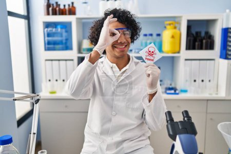 Foto de Hombre hispano con el pelo rizado trabajando en laboratorio científico sosteniendo banner tóxico sonriendo feliz haciendo signo de ok con la mano en el ojo mirando a través de los dedos - Imagen libre de derechos