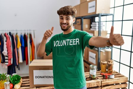 Foto de Young arab man wearing volunteer t shirt at donations stand approving doing positive gesture with hand, thumbs up smiling and happy for success. winner gesture. - Imagen libre de derechos
