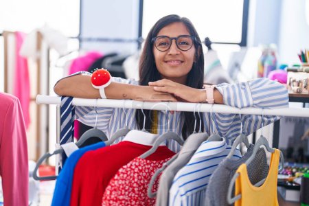 Photo for Young beautiful hispanic woman tailor smiling confident leaning on clothes rack at clothing factory - Royalty Free Image
