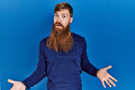 Téléchargez les photos : Homme rousse à longue barbe portant un pull bleu décontracté sur fond bleu souriant gai avec les bras ouverts comme accueil amical, salutations positives et confiantes - en image libre de droit