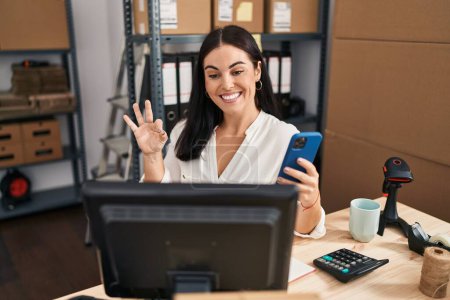 Foto de Mujer hispana joven que trabaja en el comercio electrónico de pequeñas empresas con teléfono inteligente haciendo signo bien con los dedos, sonriendo gesto amistoso excelente símbolo - Imagen libre de derechos