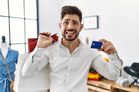 Photo for Young man with beard at retail shop holding shopping bags and credit card clueless and confused expression. doubt concept. - Royalty Free Image