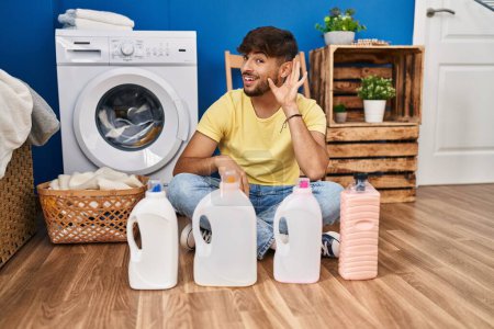 Téléchargez les photos : Arab man with beard doing laundry sitting on the floor with detergent bottle smiling with hand over ear listening and hearing to rumor or gossip. deafness concept. - en image libre de droit
