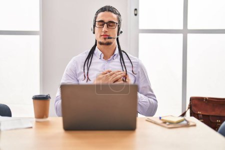 Téléchargez les photos : Homme hispanique aux cheveux longs travaillant à l'aide d'un ordinateur portable souriant avec les mains sur la poitrine, les yeux fermés avec geste reconnaissant sur le visage. concept de santé. - en image libre de droit