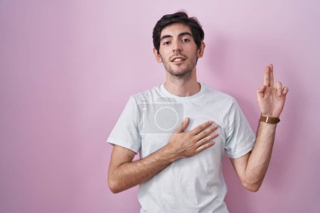 Téléchargez les photos : Jeune homme hispanique debout sur fond rose souriant jurer avec la main sur la poitrine et les doigts vers le haut, faisant un serment de fidélité promesse - en image libre de droit